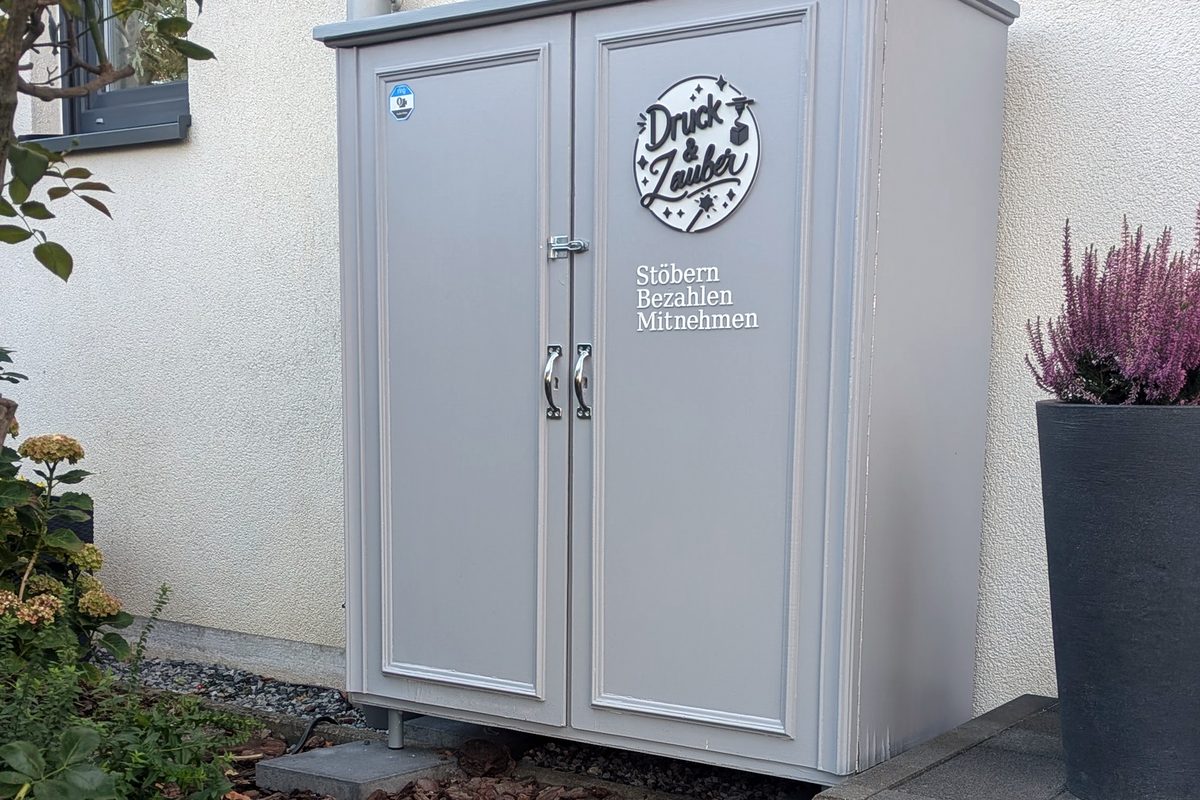 Die graue Druck-und-Zauber-Box mit Logo und dem Schriftzug „Stöbern, Bezahlen, Mitnehmen“ an der Hauswand in der Flämischen Allee 62, Viersen