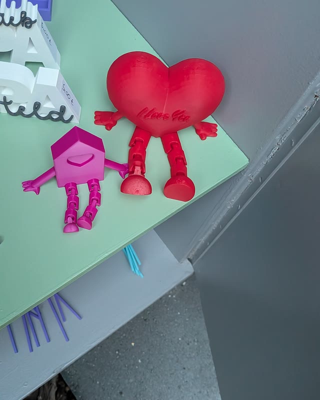 Rotes Herz mit Beinen und rosa Haus-Figur, dahinter Regenbogen-Schriftzüge