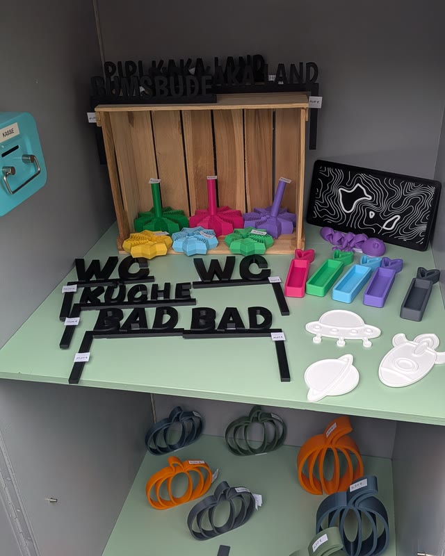 Schilder „WC“, „Küche“ und „Bad“ vor einer Holzkiste mit bunten Figuren